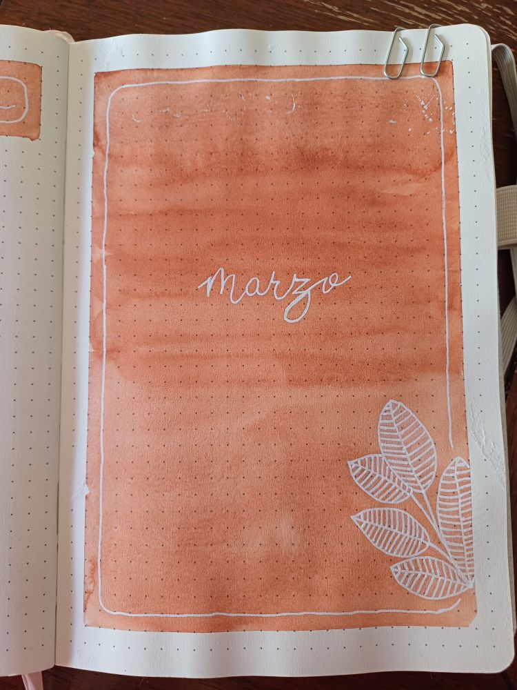 Hoja pintada de marrón con acuarela, tiene los bordes en blanco. En ella se ve una planta dibujada apareciendo por un lado y la palabra marzo escrita en letra cursiva en el centro
