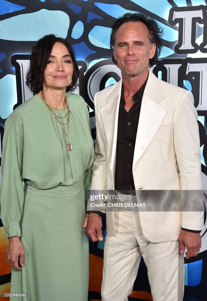 Walton Goggins and his wife Nadia standing together. Walton wears a whitr suut and black jacket. Nadia is wearing a foam green long sleeve dress.