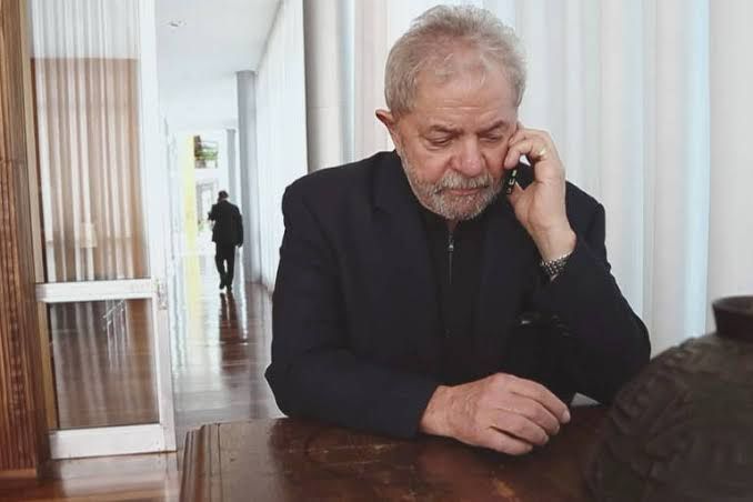 Lula ao telefone celular, homem branco sentado à mesa no palácio do planalto usando um terno preto e segurando um celular muito pequeno na mão dele 