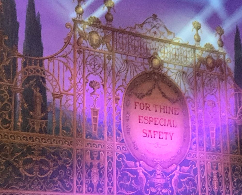 A close up (because I didn’t want to include the faces of people who might not want to be in it) of the safety curtain during the interval. The curtain is decorated to look like a huge, ornate, gold-coloured fence. A large oval in the middle has the words “FOR THINE ESPECIAL SAFETY” inside. There is ornamentation on top of ornamentation all over the fence. Winged figures, and vases, and just every twiddly over the top thing you can think of. The opposite of a fence made out of straight metal bars. 