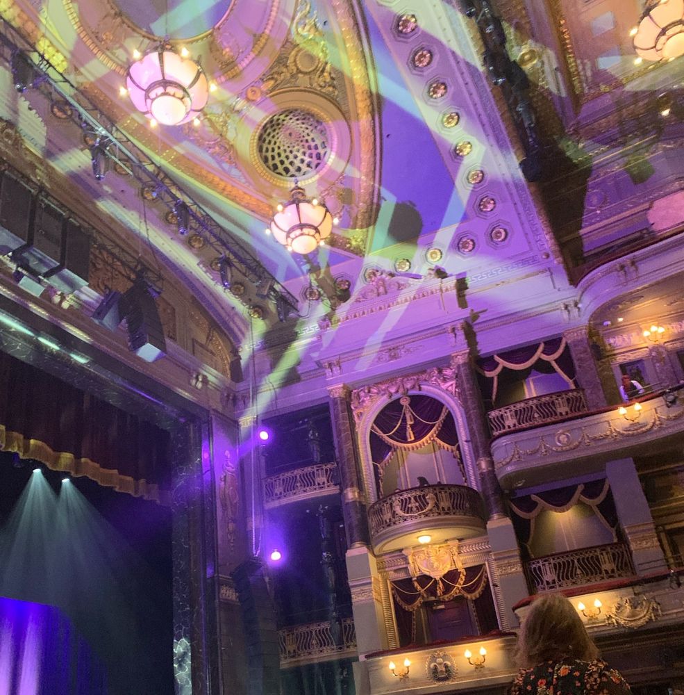 The auditorium of the Theatre Royal Drury Lane. Facing upwards from a seat in the Stalls to take a picture of a bit of the stage and its curtains, the ceiling and the boxes. Purple and gold lights are shining. There is an amount of red velvet and gilt that would be obnoxious if it wasn’t a theatre. As it is a theatre, it looks dramatic, and old-fashioned, and like there’s a non-zero chance of The Opera Ghost dropping a chandelier on someone. 