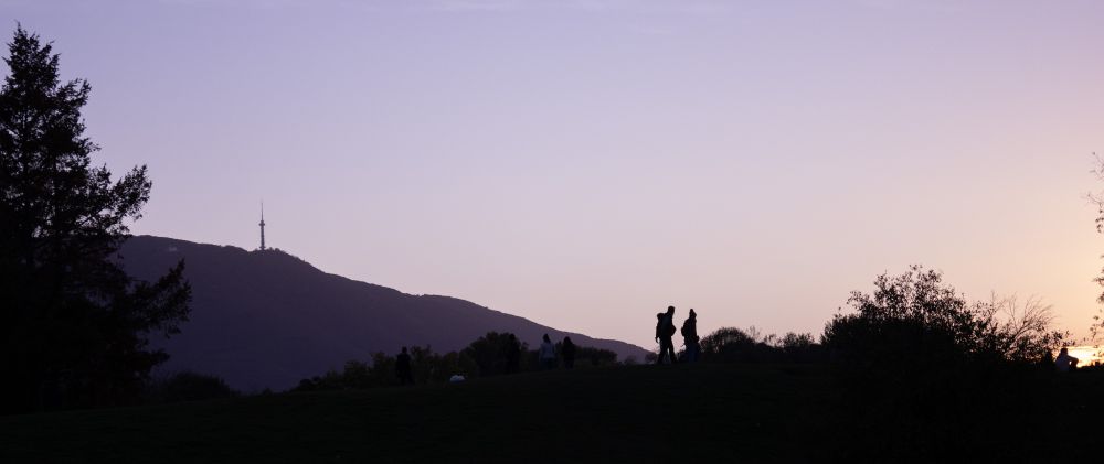 A sunset picture with the mountains at the background and 2 people and a tree in the foreground