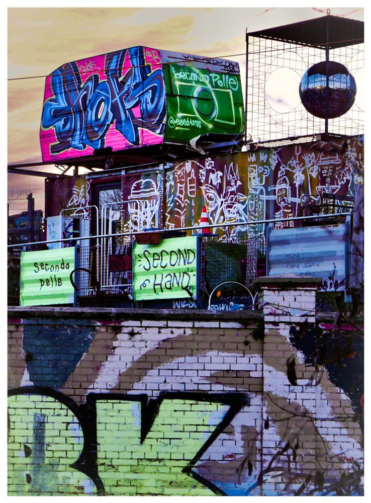 Blick über eine Mit Graffiti besprühte Ziegelmauer, dahinter gestapelte Container und oben auf ein ausrangierter Wohnwagen und eine kugelförmige Kunstinstallation. Alles mit Graffiti verziert. Der Himmel ist vom Morgenlicht gelb-orange gefärbt.