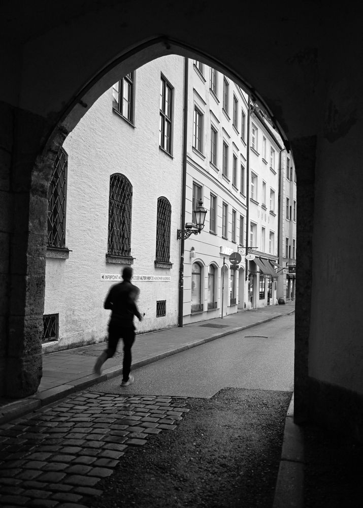 S/W-Foto. Blick durch einen Torbogen in eine historische Häuserzeile. Ein Jogger läuft die Straße entlang.