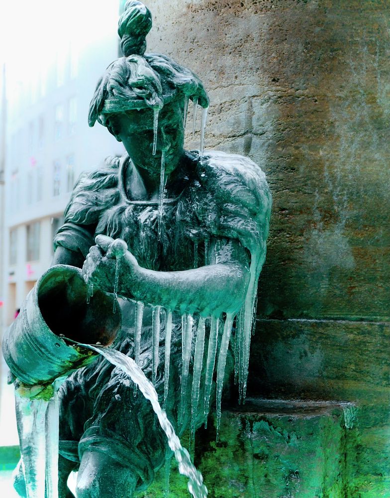 Eine metallene Brunnenfigur, die  mit Grünspan überzogen ist. Die Figur hält einen Eimer, aus dem Wasser fließt. Durch die frostigen Temperatur haben sich an der Figur mehrere Eiszapfen gebildet. Das Brunnenwasser reflektiert zusätzlich grün.