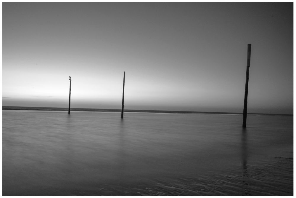 S/W-Foto.  Blick aufs Wattenmeer mit drei Leitpfosten bei Sonnenuntergang. Durch die Langzeitbelichtung ist die Wasseroberfläche seidenmatt.