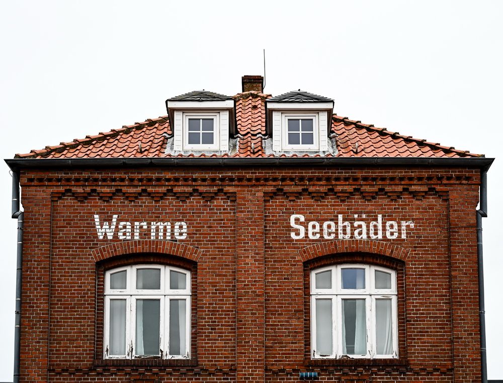 Fassadenansicht des Obergeschosses und des Dachgeschosses eines historischen Backsteingebäudes. Darauf ist mit weißer Farbe „Warme Seebäder“ geschrieben.