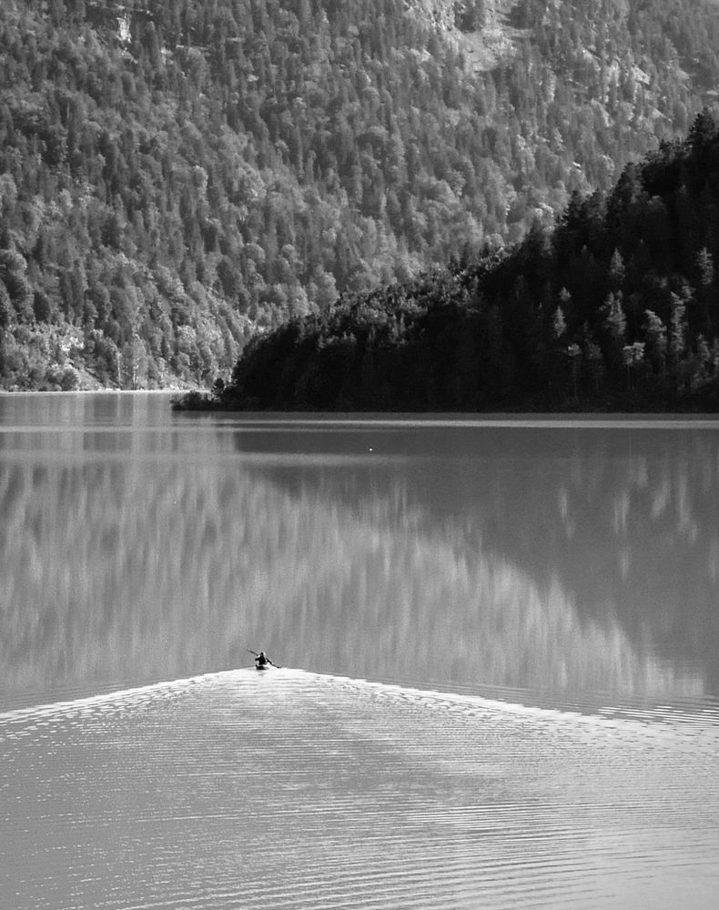 S/W-Aufnahme eines Bergsees mit glatter Wasseroberfläche, in der sich die umliegenden Berghänge spiegeln. Sehr klein ein einsamer Kanufahrer, der eine Bugwelle erzeugt.