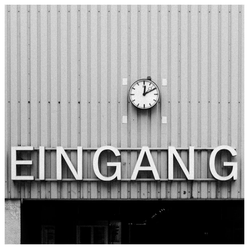 Körniges S/W-Foto eines  Schriftzugs „Eingang“ auf einer Blechfassade, darüber eine Bahnhofsuhr.