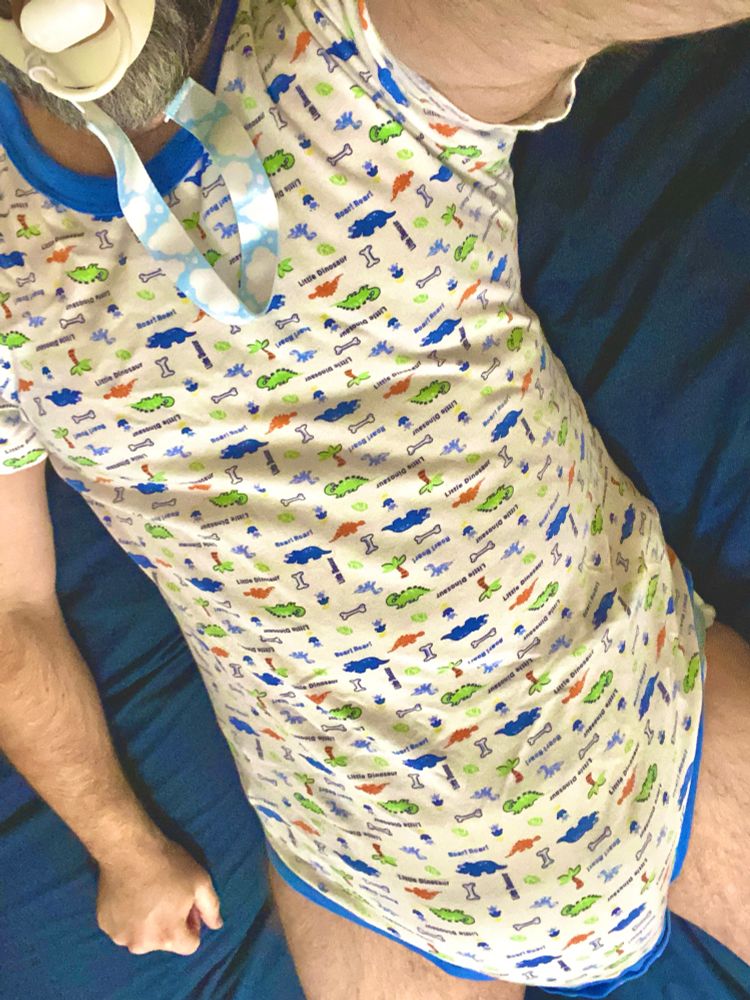 Man laying in bed in a onesie with cute dinosaurs sucking on an adult sized pacifier. The pacifier is attached to a blue sky with white clouds paci clip and is attached to the onesie at the neck. 