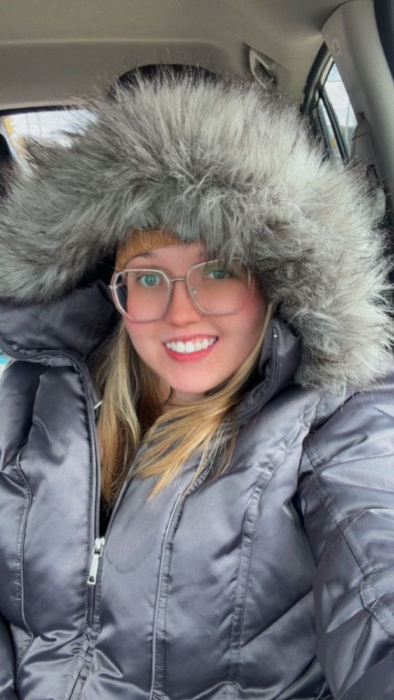 Me wearing my dark gray winter jacket with the furry gray-brown rinned hood pulled up to cover my beanie. I’m smiling despite the cold and my blonde hair is showing 