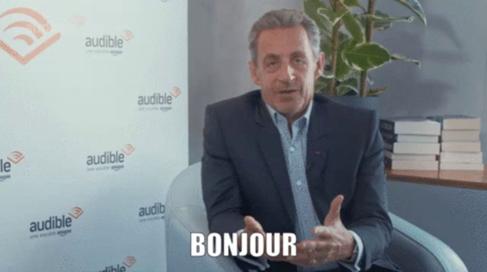 a man in a suit says bonjour in front of a wall with audible logos