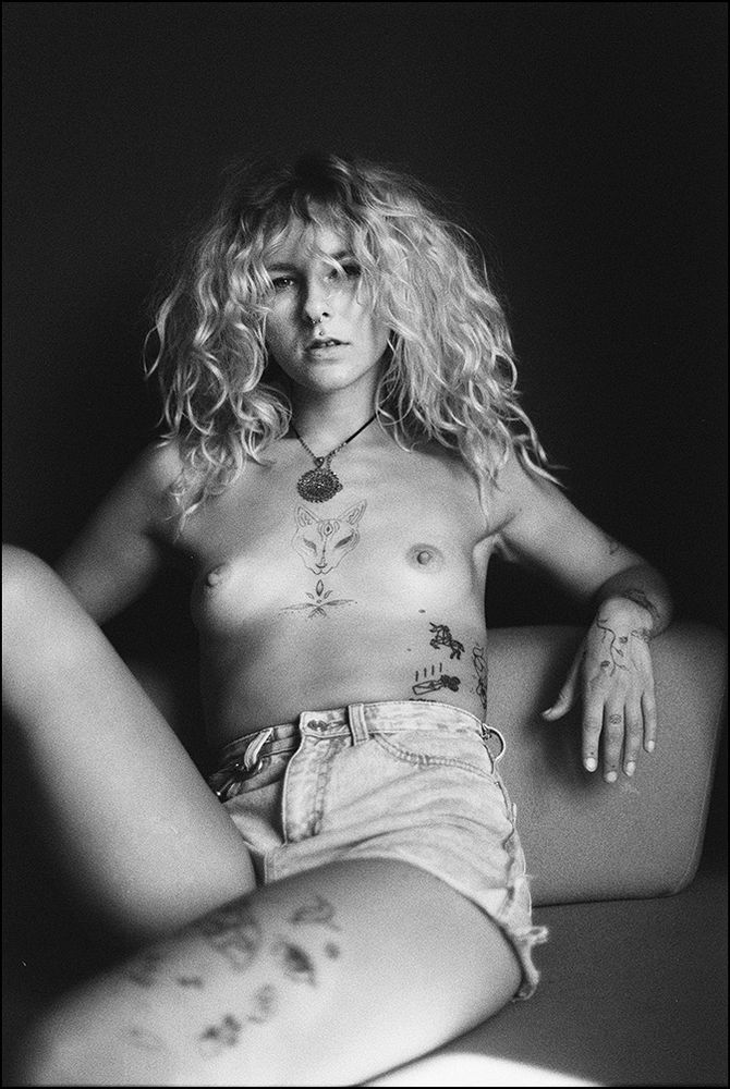 Das Schwarz-Weiß-Foto zeigt eine junge Frau mit langen, lockigen blonden Haaren, die oben ohne auf einer Couch sitzt und eine zerrissene Jeans-Shorts trägt.  Sie hat mehrere kleine Tattoos auf ihrem Oberkörper und Beinen, darunter ein Fuchs auf ihrer Brust. Ihr Blick ist direkt in die Kamera gerichtet; ihr Ausdruck ist neutral bis leicht melancholisch.  Die Komposition ist einfach, aber effektiv, mit dem Fokus auf dem Modell.
