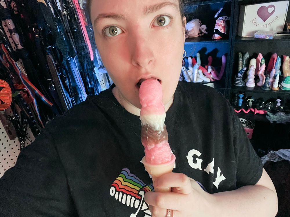 Cute chubby wife Violet facing the camera fully clothed with a silicone ice cream cone dildo pressed up to her open lips. Behind her is a huge amount of sex toys and kink gear on shelves and hanging on a peg board. 