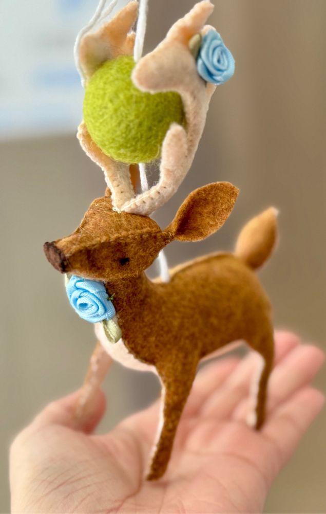 Papa deer made from brown and tan felt with a blue flower on the front of his neck and two on his tan antlers. He has a cute brown nose and black eyes. His legs and head are poseable, but he’s just standin’ there holding the light green wool ball between his antlers. 