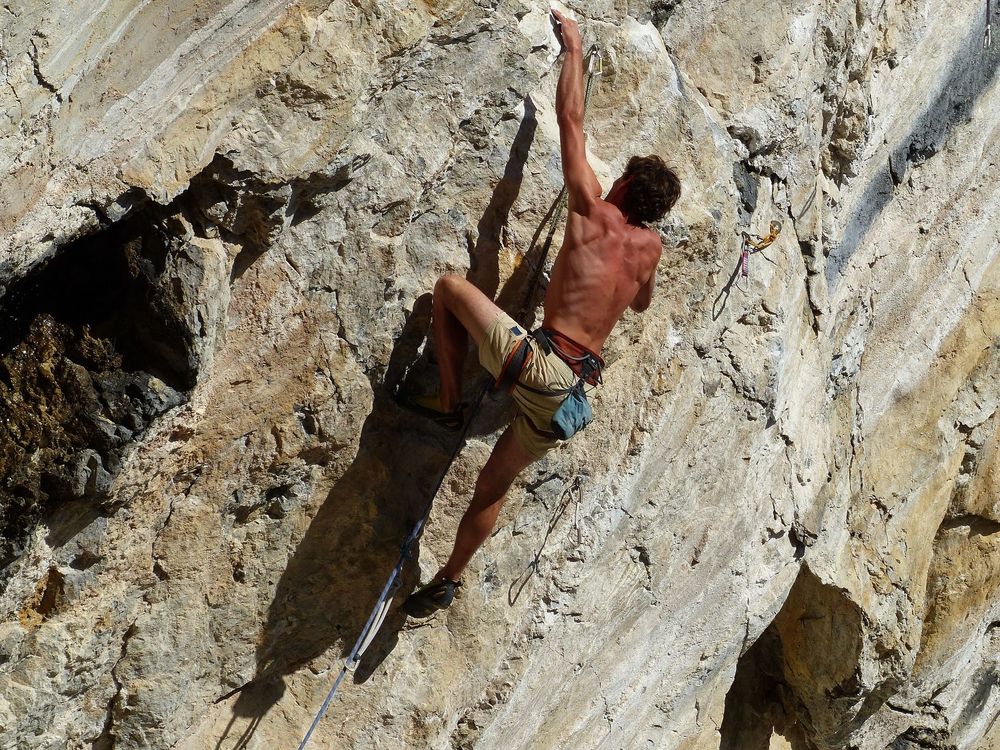 A lean, muscular shirtless man scales a sheer cliff face with his bare hands.