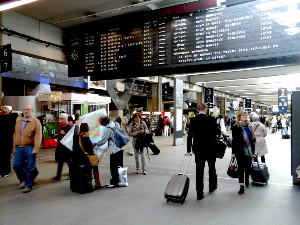 voyageurs et voyageuses dans la gare montparnasse, sous le panneau d'affichage ancien, en 2014. Deux personnes consultent curieusement un planisphère du monde...