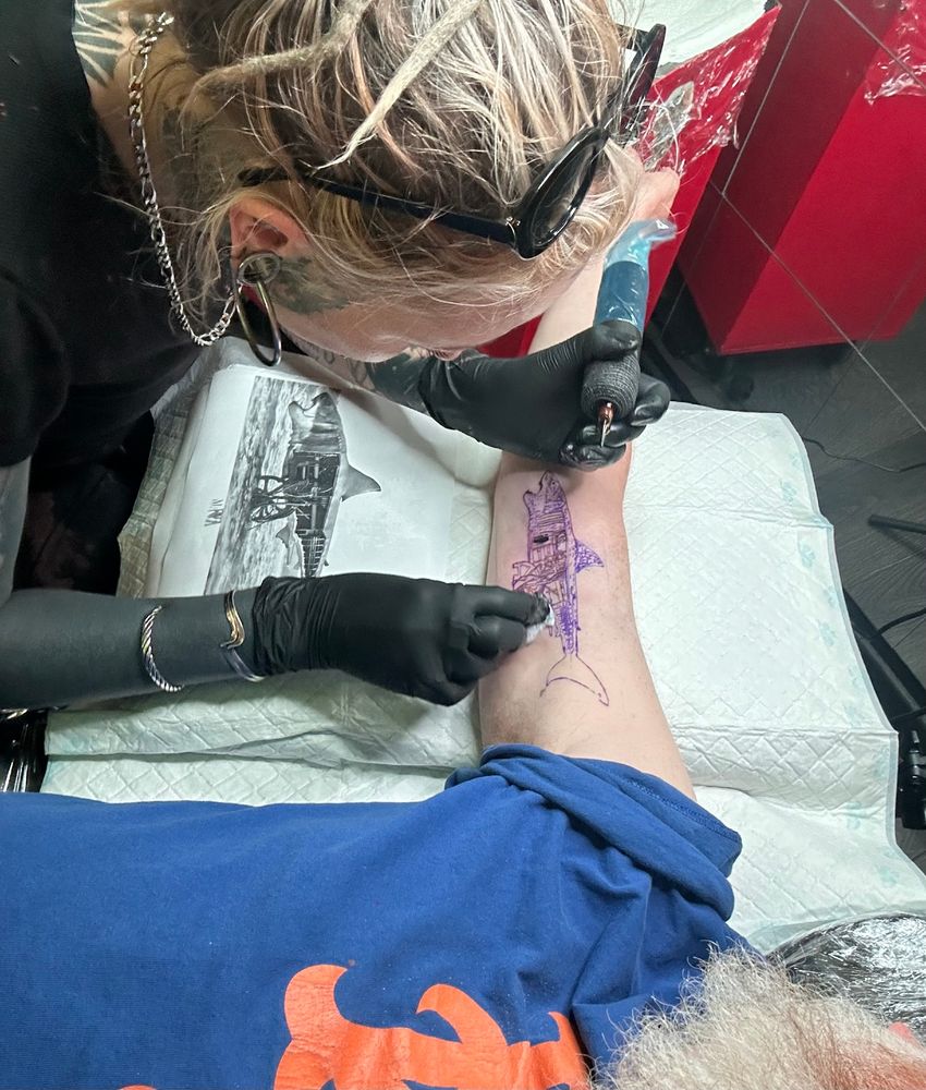 Blonde woman with dreads tattoos white man's inside bicep using a stencil