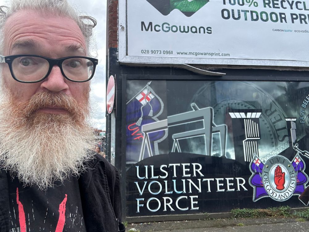 A bearded man looking freaked out beside a big mural depicting the UVF, a murderous terrorist organisation proscribed by the UK's government