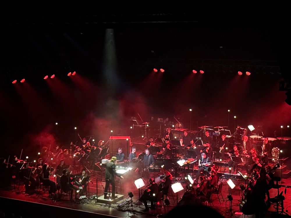 A stage populated by an orchestra and members of Sigur Ros. The lights are red, with a white spotlight shining straight down on the conductor. 