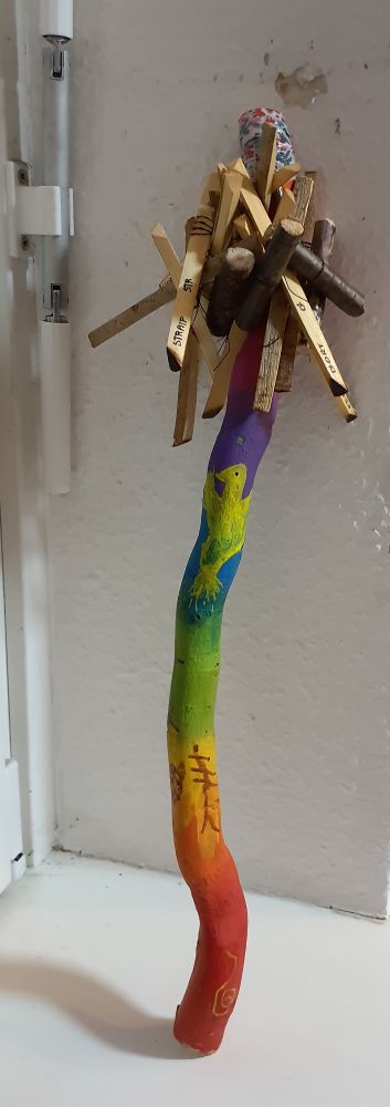 Foto of a rattle painted in rainbow colours with the symbol of a bear, a phoenix and tow ogham symbols on it and some sticks hanging from it that make the sound. ü