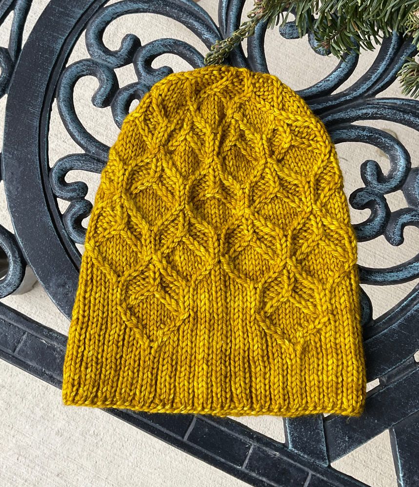 A yellow-gold hat that uses crossed stitches and 1/1 cables to replicate the cells in honeycomb. The photo is taken outside on a metal bench on a concrete patio. 