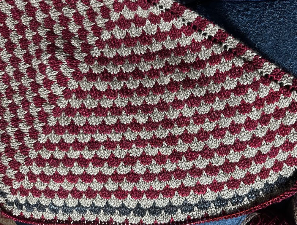 A partial view of a knitting shawl that uses a dip stitch to go into rows below to allude to a keel/keel centerboard of a boat. There are alternating rows of gray and red. A repeat of blue rows will be followed but gray, red, and gray before appearing again. Pattern by Isabell Kraemer