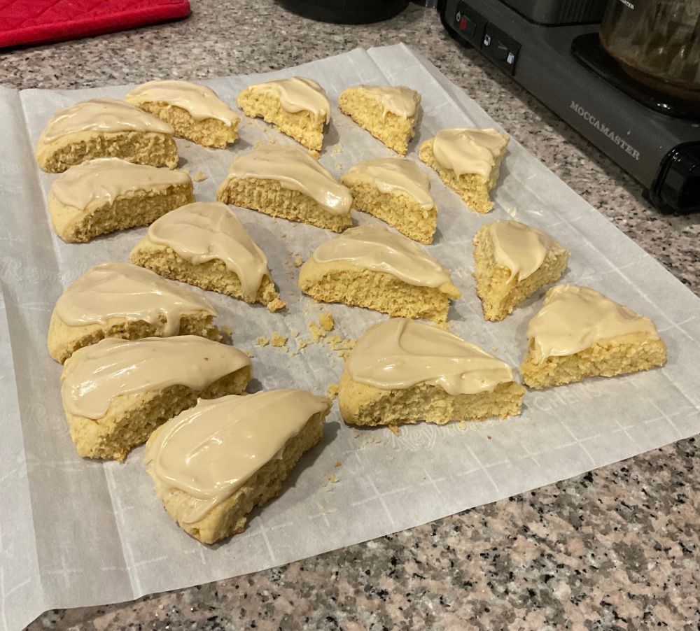 Sixteen frosted maple scones - on a sheet of parchment paper they were baked on - sitting on a granite countertop. 