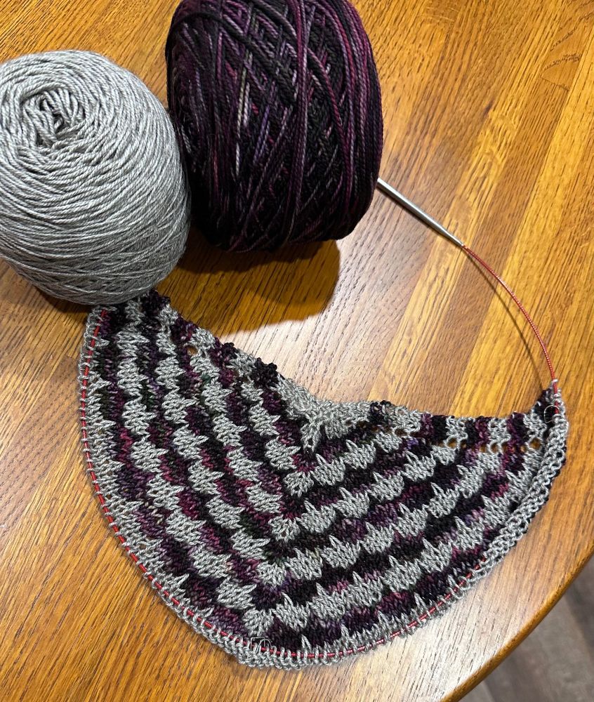 The beginning of a knitted shawl on a wooden table. The pattern is a 4-row repeat with a dip stitch, reaching down into earlier knitting, at a regular repeat. The main color is a yak, silk, and merino yarn in an undyed gray. The multicolor yarn is variegated reds from almost black to rose and gray. 