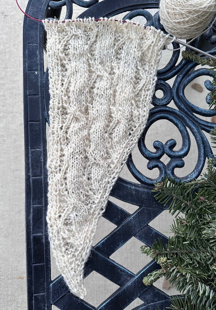 The beginning of a white scarf photographed on a metal bench. Part of a Christmas wreath along the one edge of the photo. The texture of the scarf is panels of garter stitch overlaid with left and right twists and stockinette stitch with a cable. 