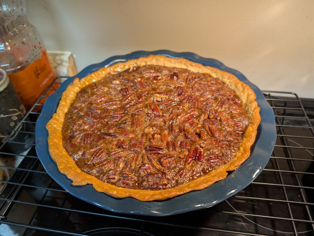 A photo of a fresh-baked pecan pie in a blue pie dish, cooling on a rack above the stove. 