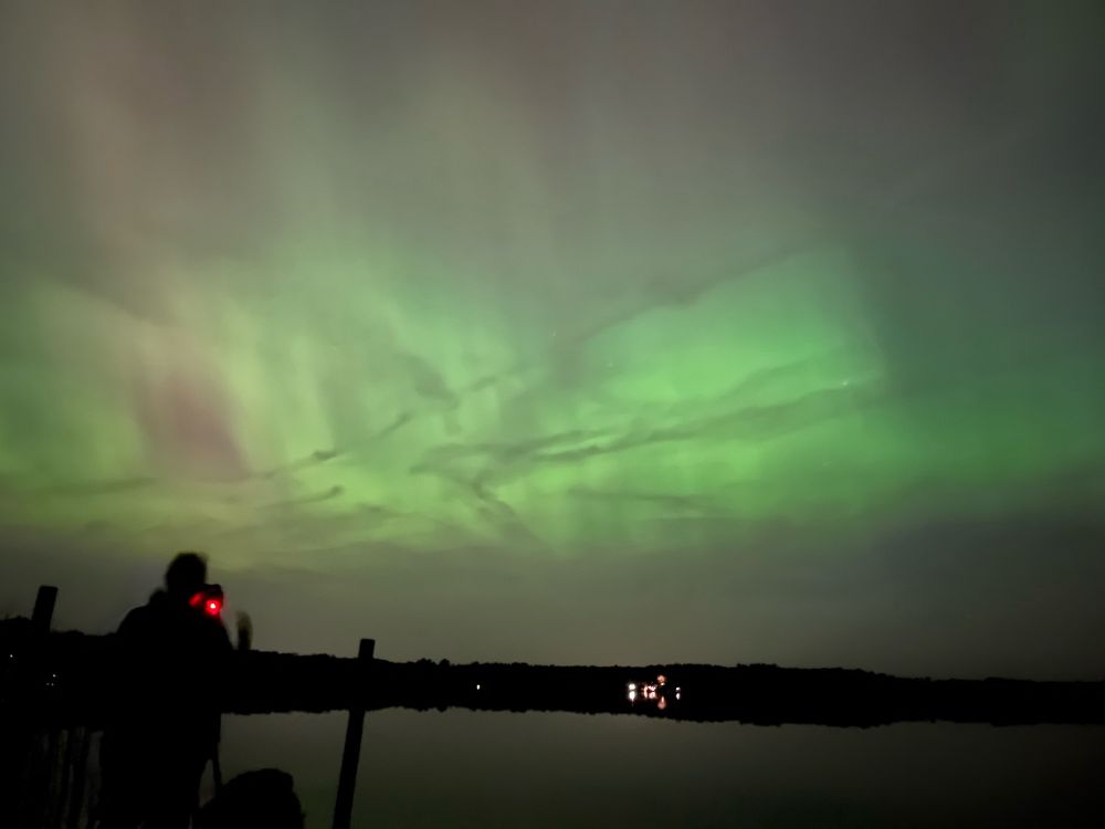 A sky full of green aurora silhouetting long clouds and contrails.
