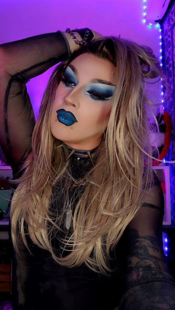 Charli Nicks magnifique en drag, perruque blonde décoiffée, haut en résille, spikes, chaînes, piercings, maquillage bleu et noir, pose approximativement inspirée et mélancolique 
