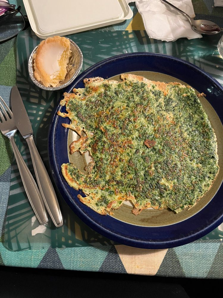 Bestick till vänster i bild. En mörkblå tallrik står på en grönmönstrad plasttablett på en bordsduk med mönster av trianglar i olika nyanser. På tallriken ligger en spenatpannkaka med baksidan uppåt. Uppe till vänster om tallriken ligger en aluminiumform med en halväten mazarin. Överst i bild syns en bit av locket från ett smörpaket, en bit hushållspapper och något som jag tror är skaftet av en sked