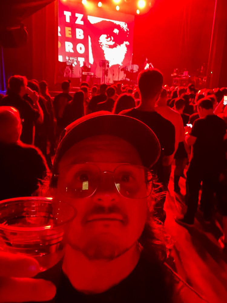 Selfie holding up cup of diet coke, I’m bathed in red light. Behind me across a crowd of people Nitzer Ebb performs on stage 