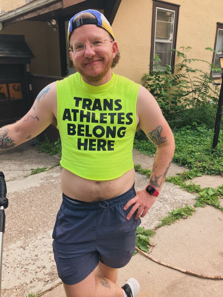 me, a white trans guy with a red beard, wearing a bright yellow crop top that says trans athletes belong here 