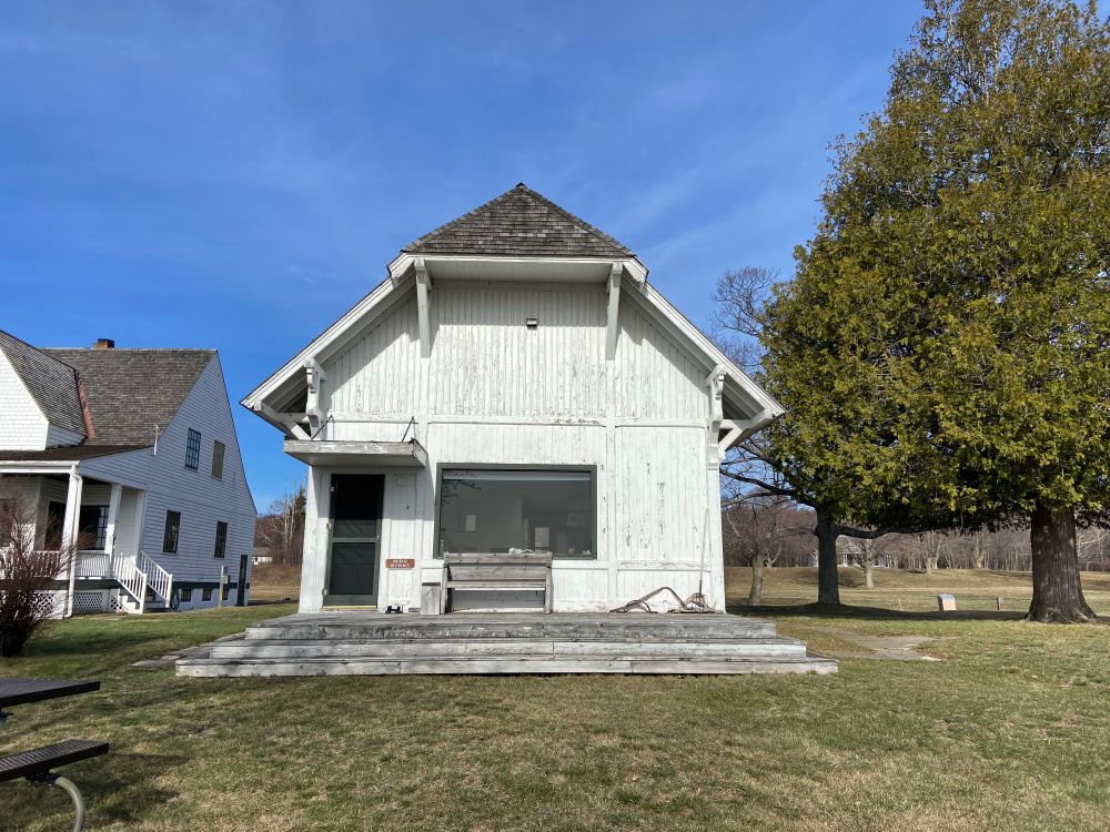 An old boathouse painted white with cedar shingles for a roof, converted into simple living quarters. A bay window looking out on the water (out of frame) stands in place of the big door they would wheel boats through. Blue sky, morning sunlight. 