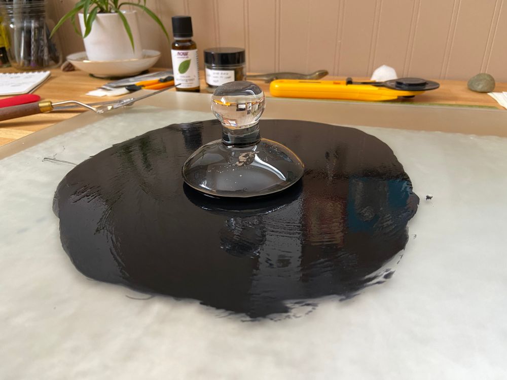 A photo of a circle of wet black ink spread on a frosted glass slab. A clear glass muller sits at the center.