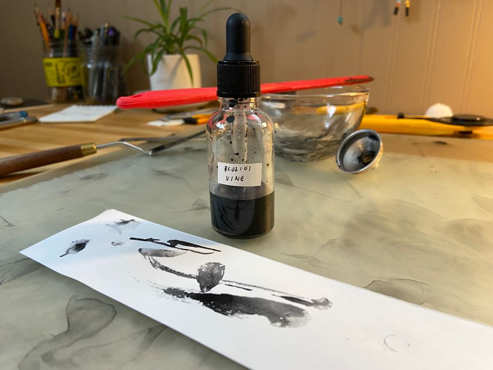 A photo of a small bottle of black ink labeled "BC02:01 VINE" sitting among ink-stained art supplies on a desk. Next to the bottle is a scrap of smooth paper with black brushstrokes.
