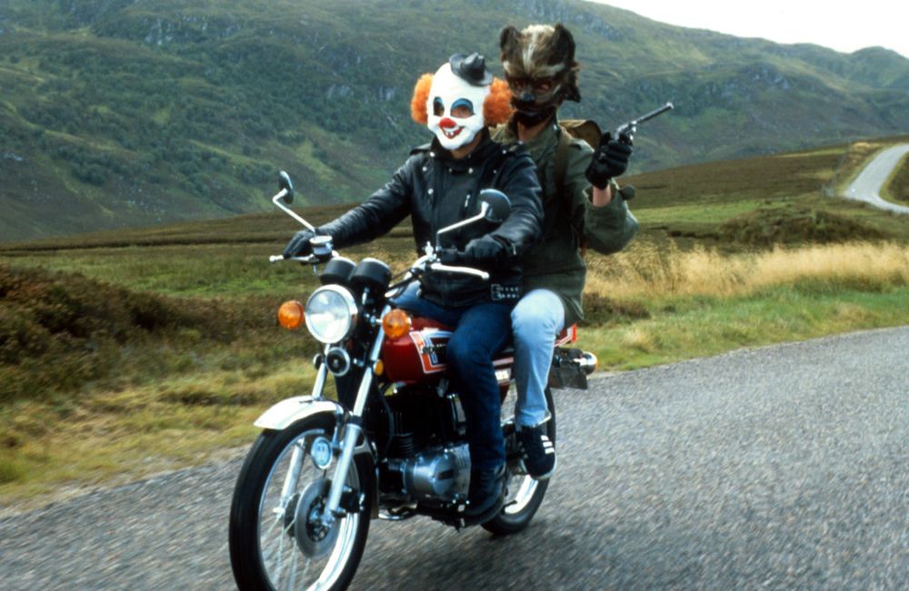 Two men on the back of a motorbike riding along a remote Scottish road. One is wearing a clown mask, the other a wolf-man mask.