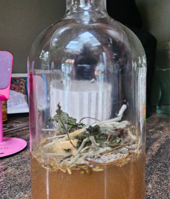a large glass bottle filled almost hallway with herbs and a light brown liquid sitting on top of a marble counter