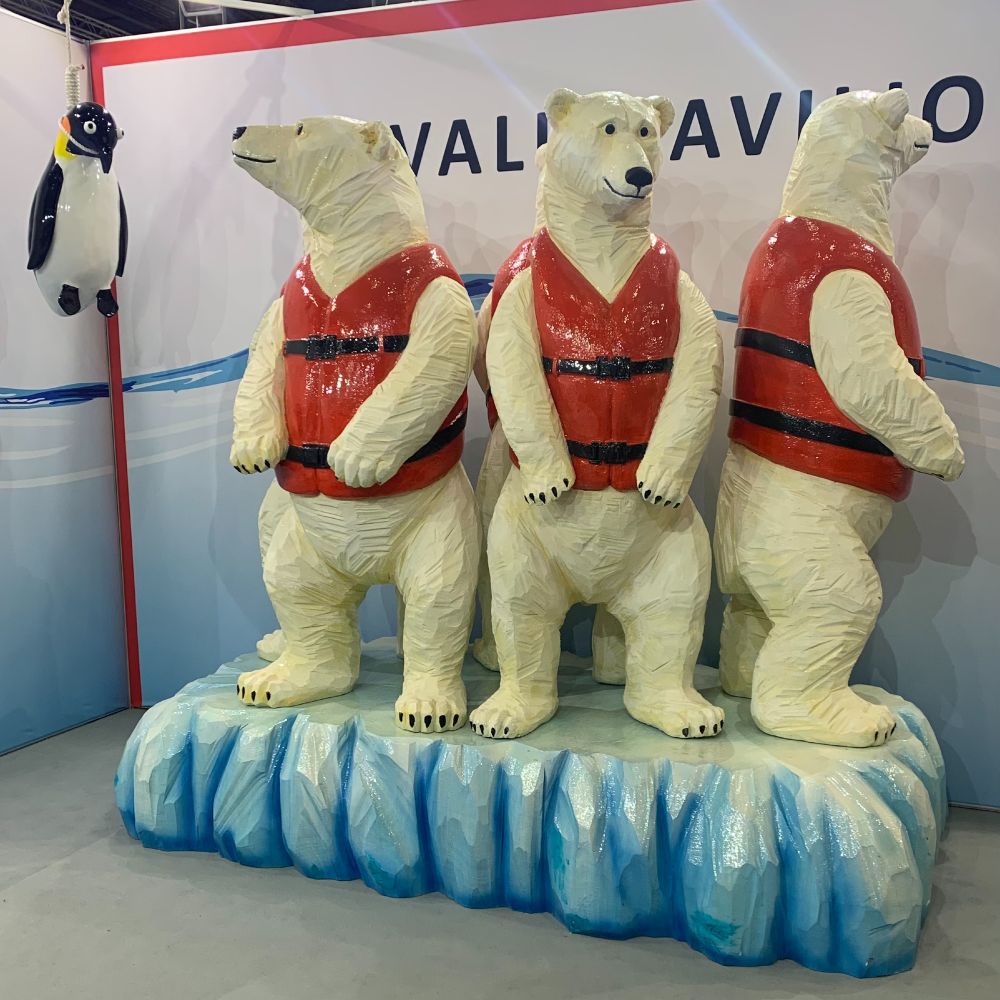 A diorama of 3 polar bears standing on a small iceberg, wearing orange lifejackets. One of them is looking at a penguin that appears to have been hanged.