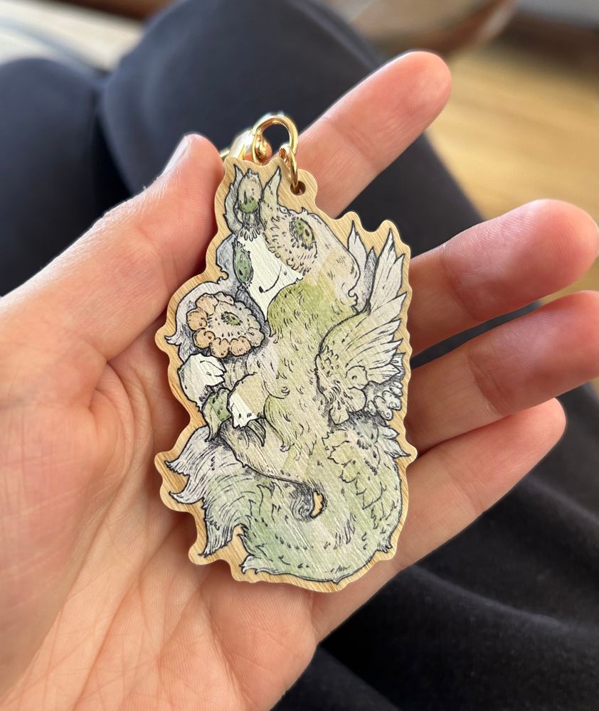 Photo of a wooden keychain in the shape of a green fluffy worm with a single eye and feathers holding a flower