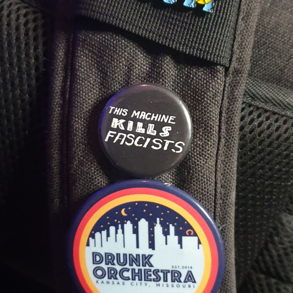 Camera bag strap prominently featuring a few buttons. The one in the center of the photo says "This machine kills fascists" in Woody Guthrie's handwriting. The one below it says "Drunk Orchestra" because I always have to give a shout out to my people.