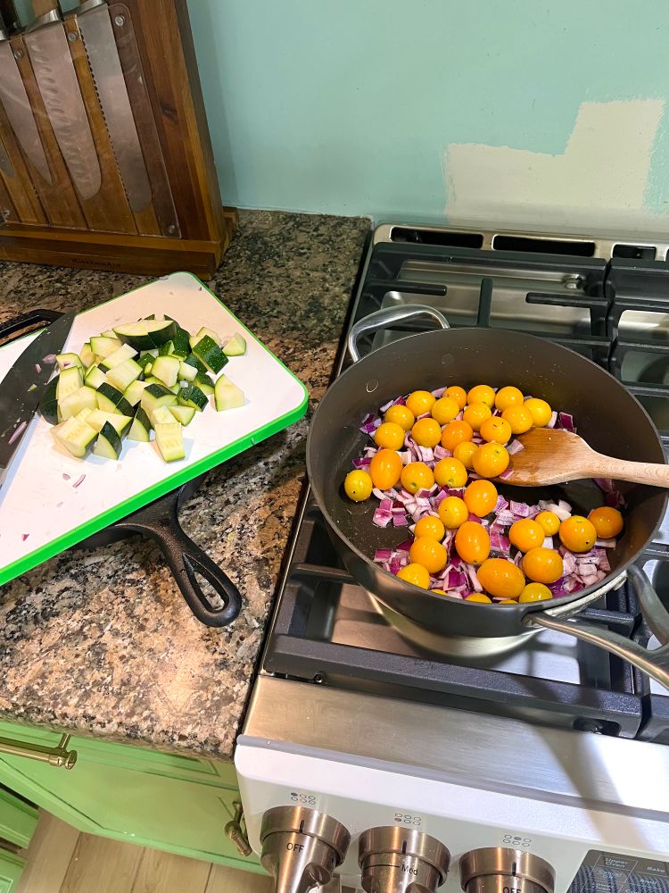 A skillet on a stove with chopped red onions and golden cherry tomatoes. Next to it on the counter is a cutting board with chopped zucchini. 