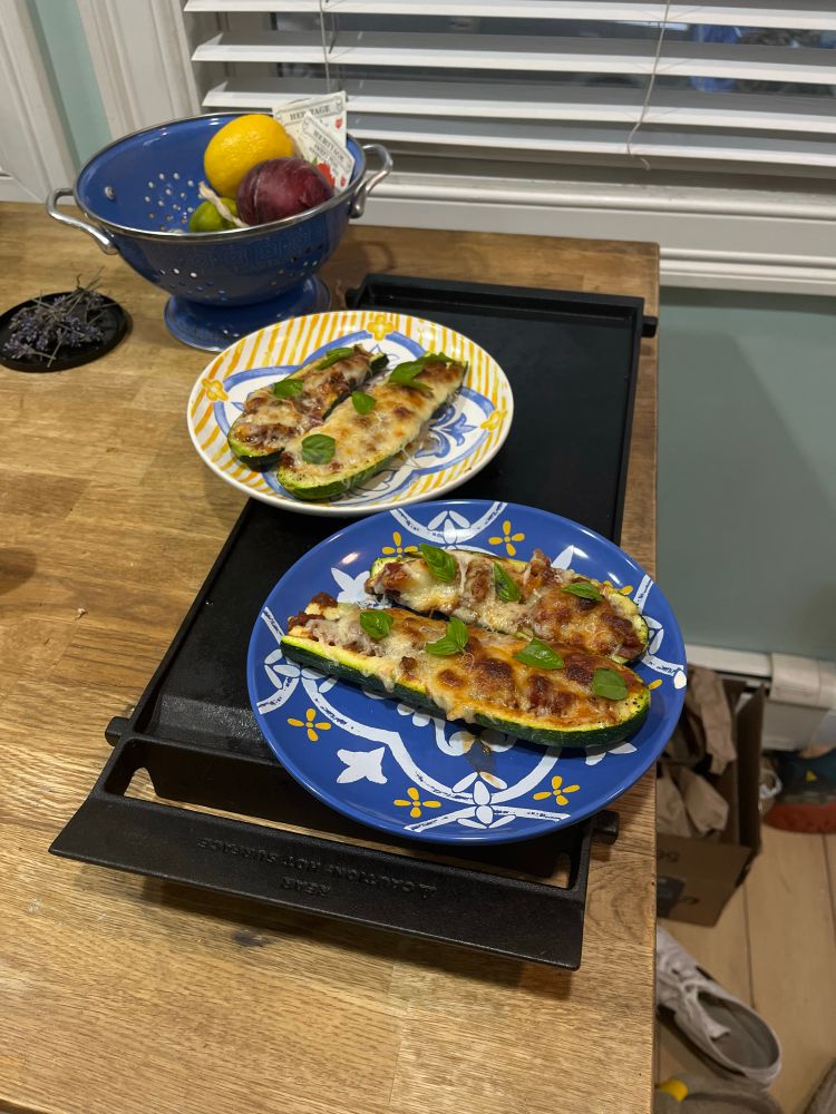 Two plates of zucchini boats on a black skillet on a wooden table. They are filled with bolognese and topped with browned cheese and basil. 