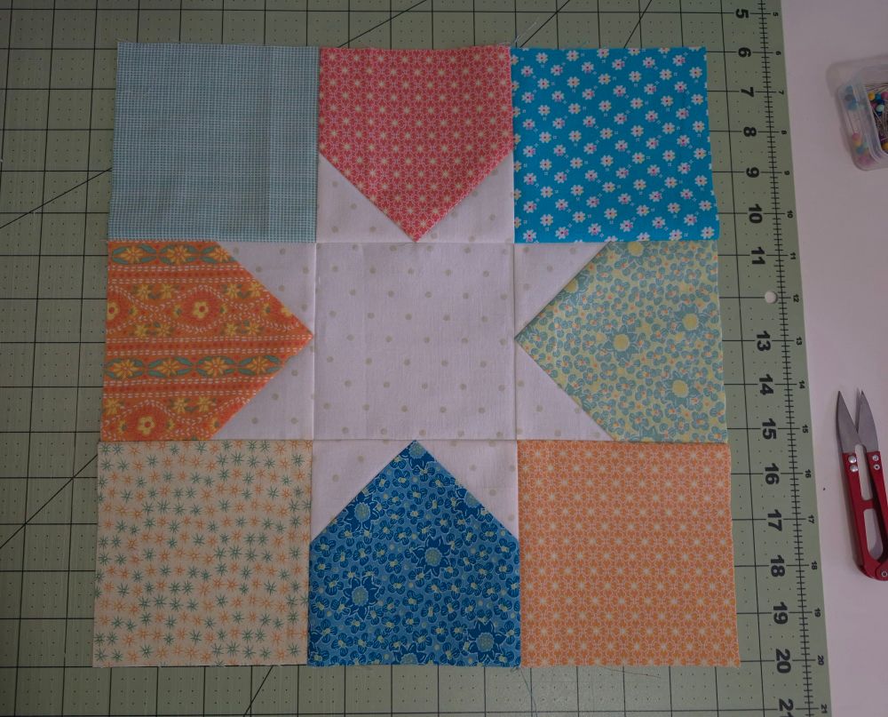 A quilt block consisting of 9 different brightly coloured squares with a star in the center is sitting on a green cutting mat. 