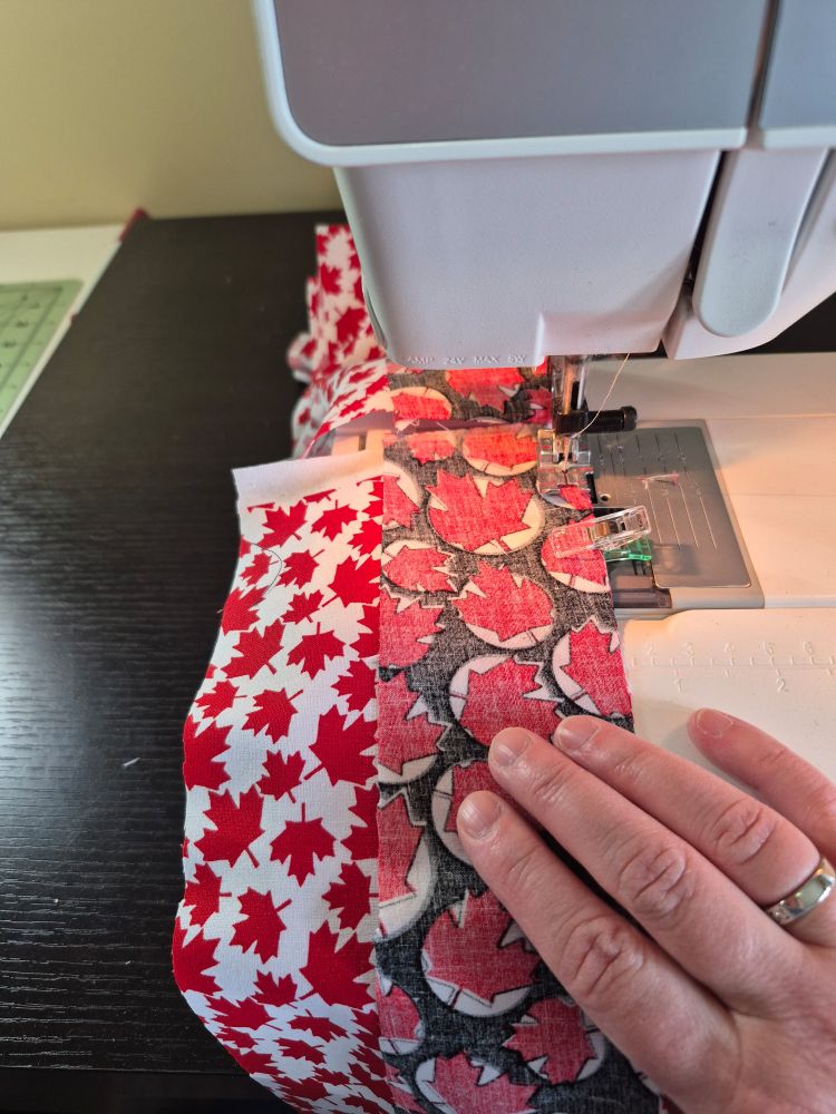 Strips of red white and black maple leaf fabric are being fed through a sewing machine. 