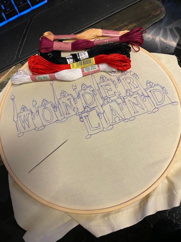 Embroidery frame with inside a fairly light yellow fabric drawn on it the KH1 Wonderland logo. With not far from white, red, black, very light orange and dark purple embroidery threads