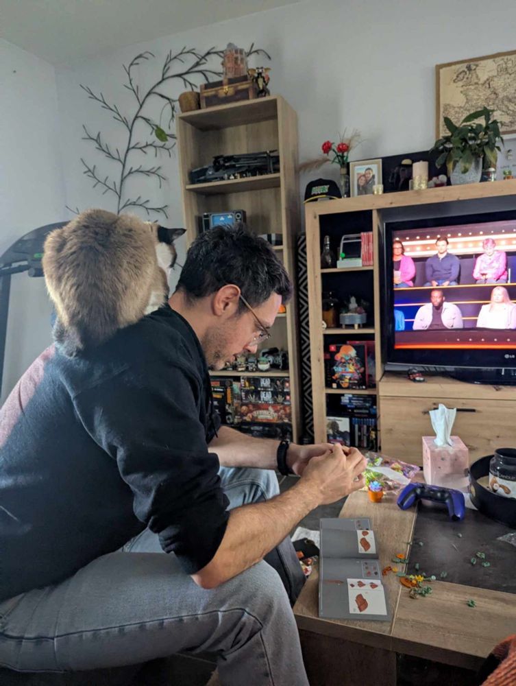 Man doing Lego in front of  TV with with a Siamese cat sitting on his back